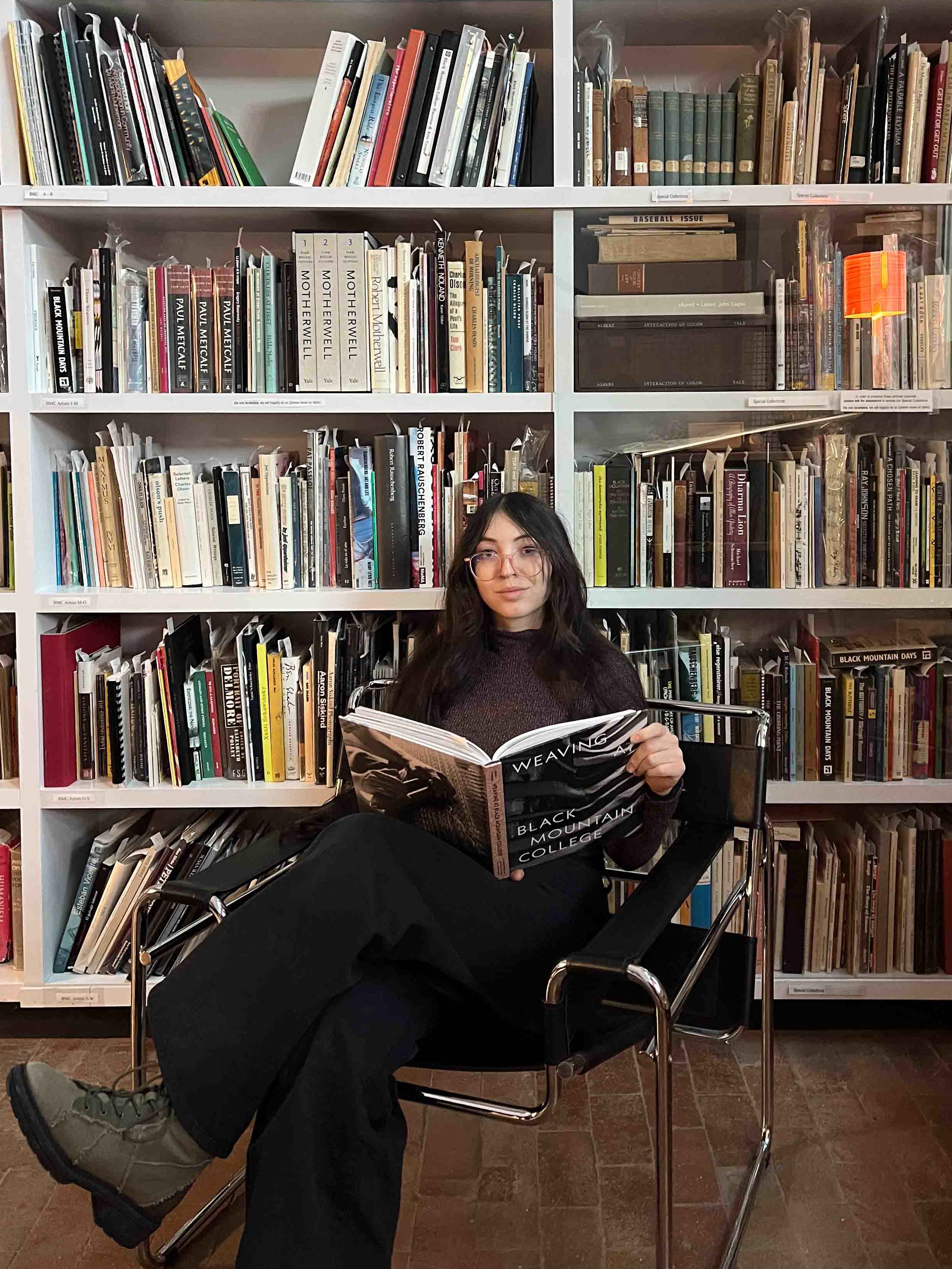 A woman sitting in a library holding a book open and looking at the camera
