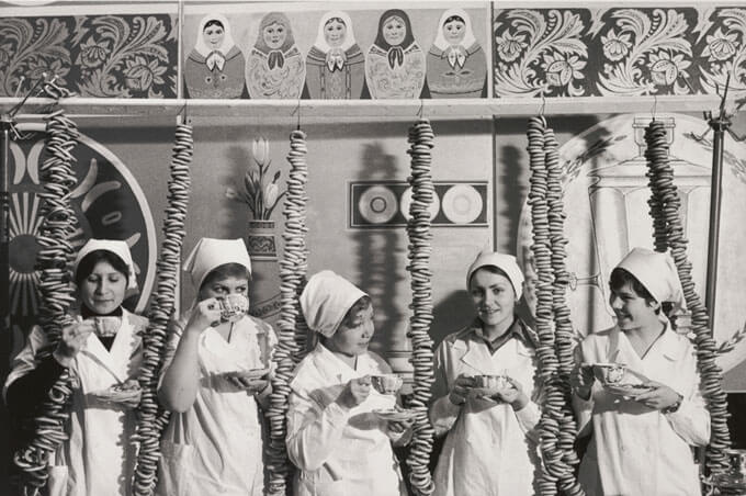 Five women in white uniforms drink tea surrounded by vertical strings of bagels and decorative Russian motifs.