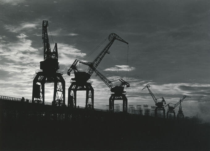 Black and white photo of construction cranes standing tall against a sunset sky.