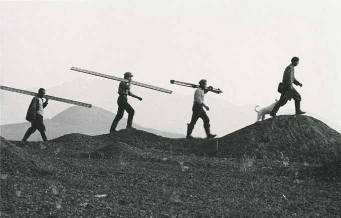 Black and white photo of three people carrying large rods over their shoulders, led by a man with a dog.