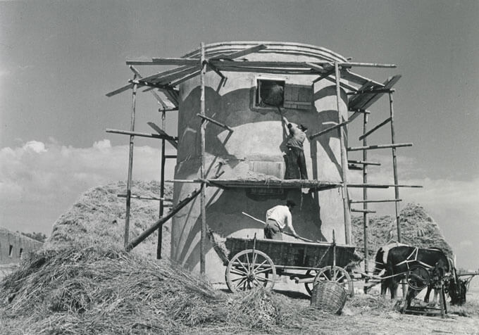 Two people transfer hay from a horse-drawn cart into a silo.