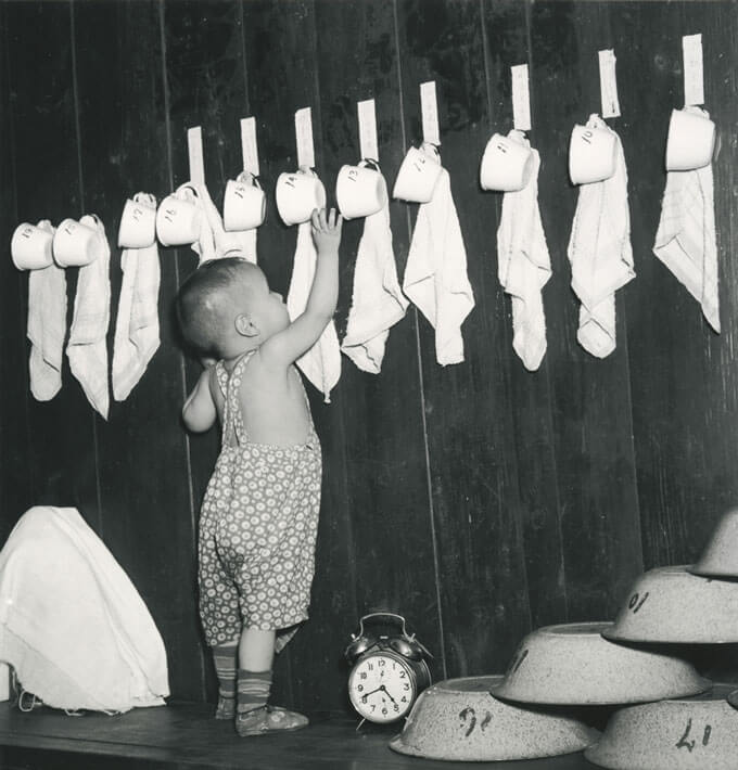 A black and white photo of a toddler reaching for a teacup hanging on the wall.