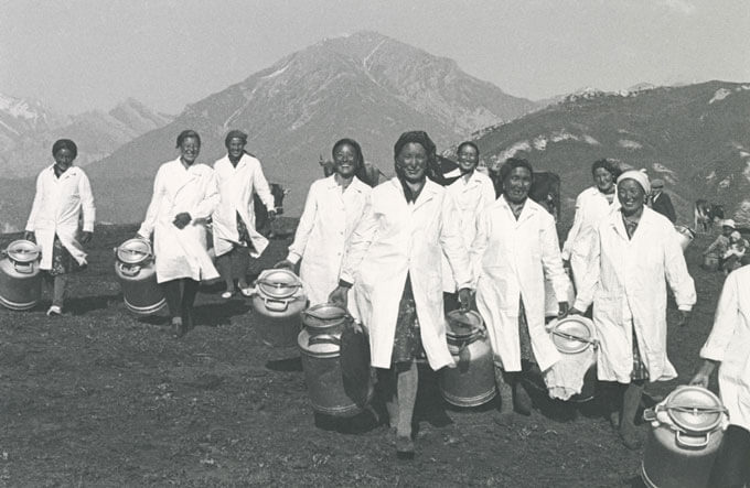A group of milkmaids in white coats carry milk jugs in a rural setting
