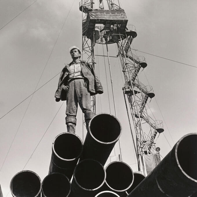 A black and white photo of an oil worker standing atop metal pipes with a tall structure in the background.