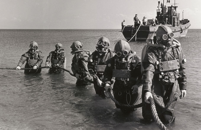 Divers in vintage deep-sea suits emerge from water, holding a rope with a ship in the background.