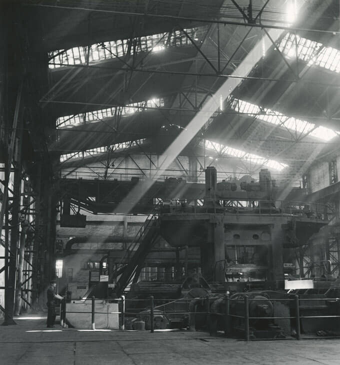 A black and white photo of a man standing in a large factory.