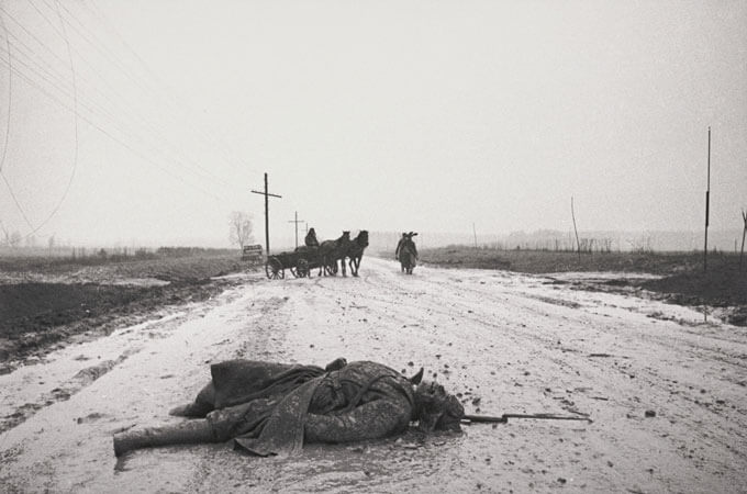 Black and white photo of a soldier lying dead on a road. A horse-drawn cart is positioned in the background.