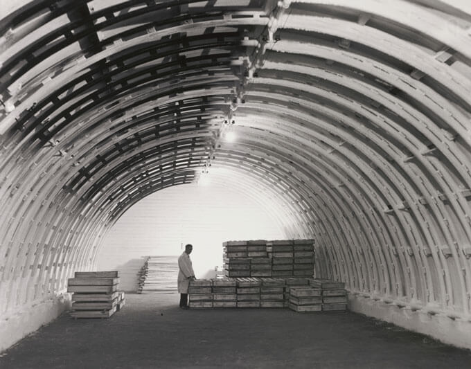 A person stands in front of stacks of wooden crates inside a storage facility with a curved roof.