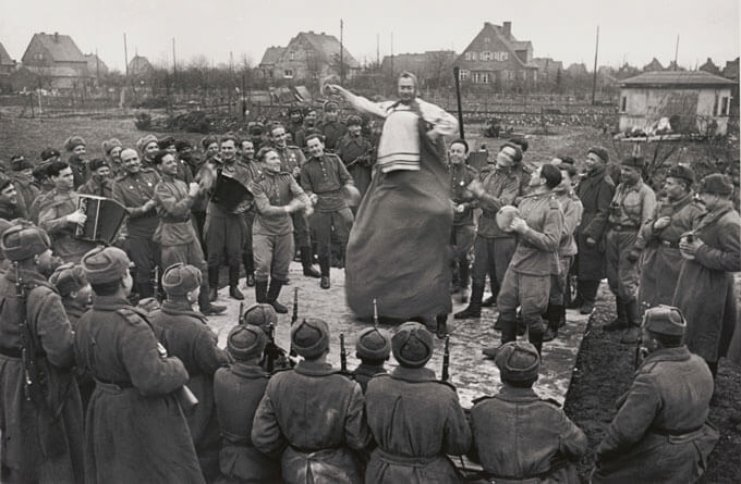 Group of soldiers gather around a man dancing in traditional attire with musical accompaniment in a rural setting.
