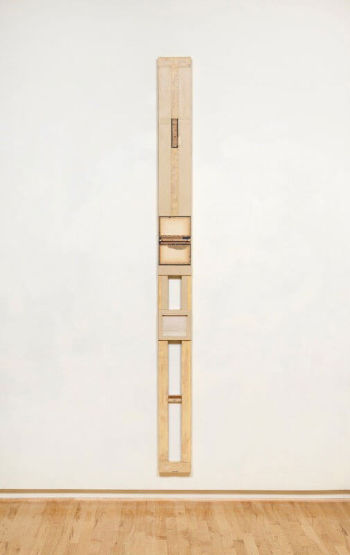 Elongated vertical wooden sculpture with multiple segments on a white wall.