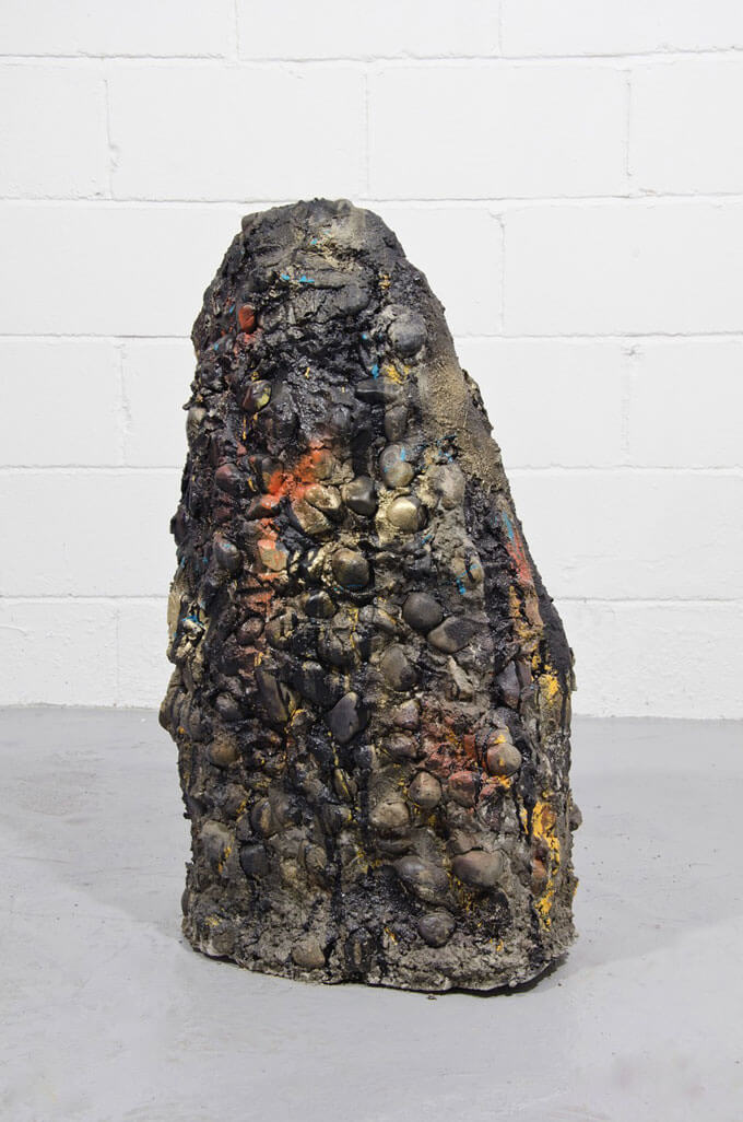 A large, textured sculpture with dark tones and colorful highlights, resembling a boulder on a grey floor against a white brick wall.