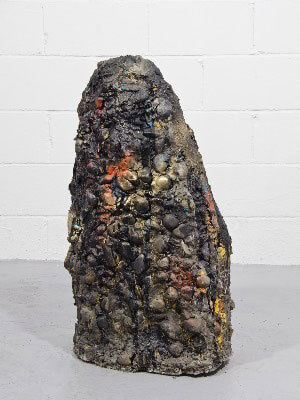 A large, textured sculpture with dark tones and colorful highlights, resembling a boulder on a grey floor against a white brick wall.