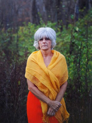 Portrait of an adult with short white hair in a red dress and yellow shawl standing in a closely cropped landscape