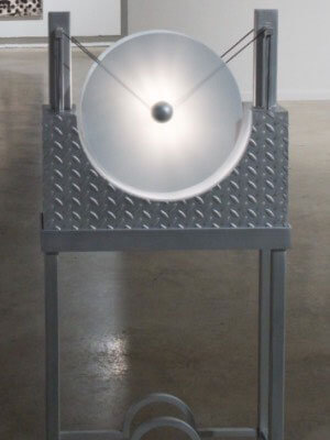 A metallic sculpture with a circular frosted disc on a diamond-patterned metal stand.