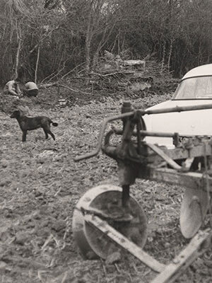 Car parked next to a plow in a field. Two men kneel over the ground in the background, with a dog standing in the foreground.