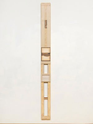 Elongated vertical wooden sculpture with multiple segments on a white wall.
