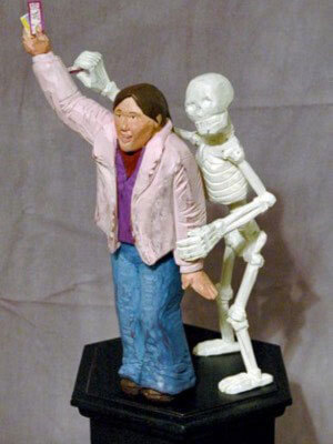 Sculpture of a skeleton standing behind an adult figure while holding their left arm down and raising their right arm up to show tickets both standing on a black base
