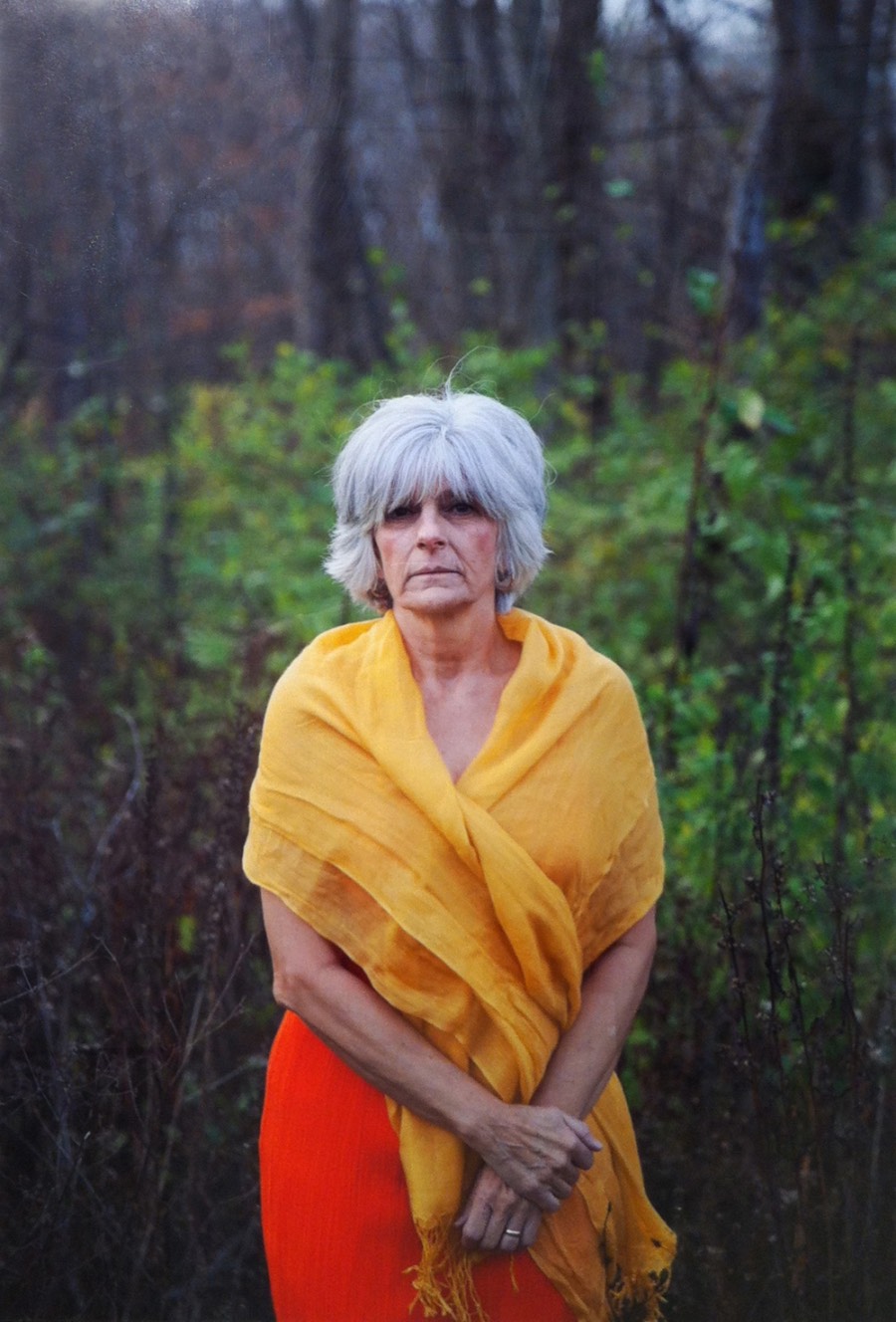Portrait of an adult with short white hair in a red dress and yellow shawl standing in a closely cropped landscape 