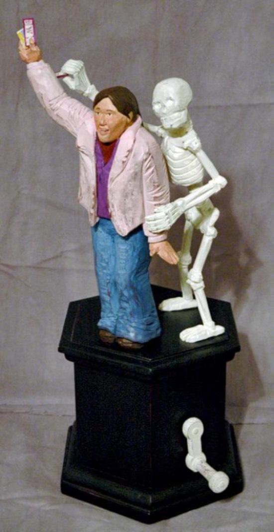 Sculpture of a skeleton standing behind an adult figure while holding their left arm down and raising their right arm up to show tickets both standing on a black base