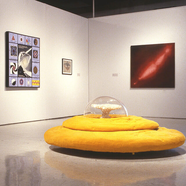 An art gallery with a yellow, circular, layered sculpture in the center, resembling a UFO with a transparent dome on top enclosing a smaller sculptural element.