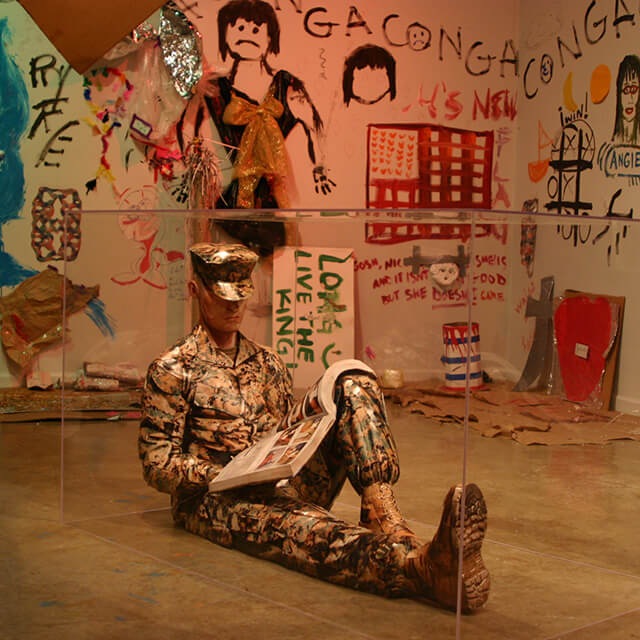 A mannequin dressed in a military camouflage uniform sitting on the floor in a cross-legged position, holding a newspaper. The mannequin is positioned inside an art installation, surrounded by walls heavily adorned with various paintings and graffiti. The artwork on the walls includes abstract figures, faces, phrases, and a mix of vibrant colors such as blue, red, green, and black. Some paintings appear to depict human figures and shapes, while others contain abstract patterns.