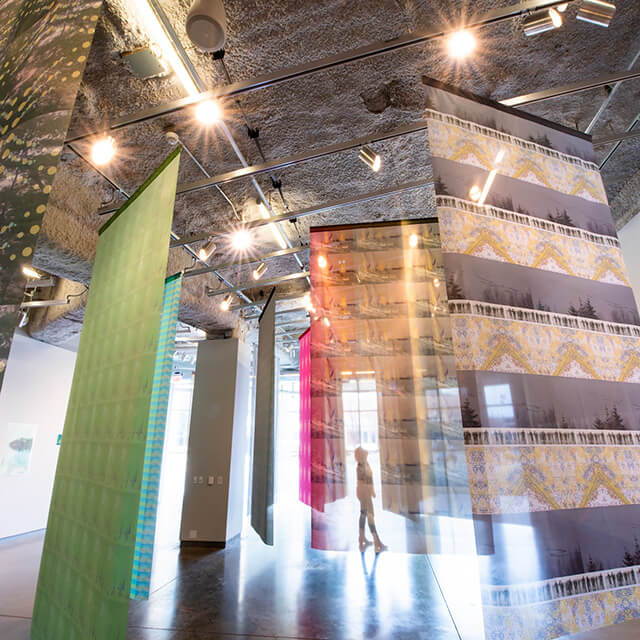 An art installation in a spacious gallery with multiple large translucent panels hanging from the ceiling. These panels display a variety of patterns and colors, including green, blue, pink, orange, and a multi-patterned yellow and gray design.