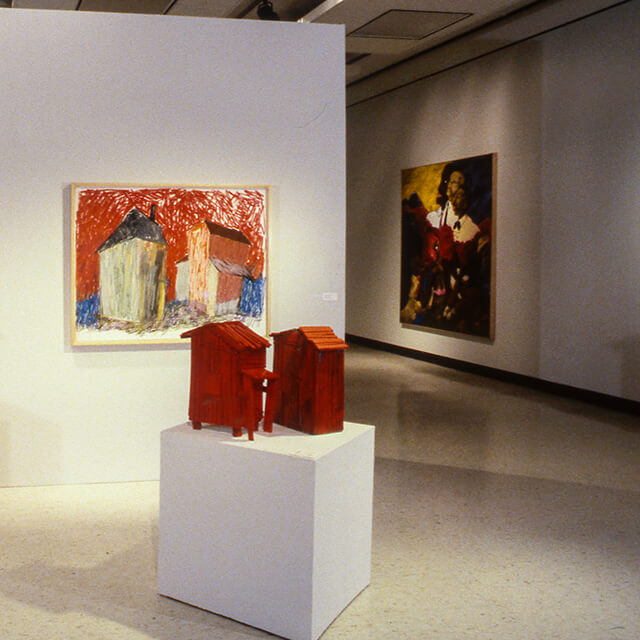 An art gallery interior– in the foreground, there are two small, red, abstract wooden sculptures on a white pedestal. The sculptures have an angular, textured design. On the wall behind the pedestal is a colorful painting of three buildings with a vibrant red and blue background. The painting has a textured, almost childlike style. Further back, an additional painting hangs on the adjoining wall, showcasing a figure against a dark background with rich, earth-toned colors.
