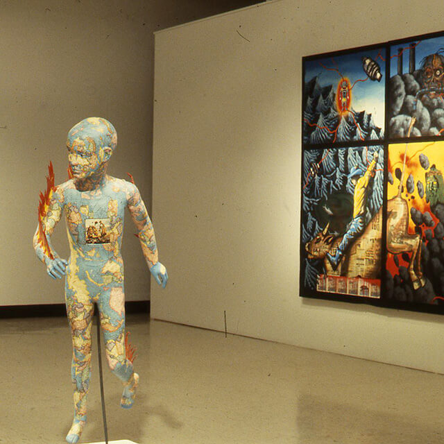 An indoor art gallery with a prominent sculpture in the foreground and a colorful painting on the wall in the background. The sculpture is of a human figure, standing upright, with a surface covered in a map-like texture and small, vivid flames emerging from its body. The figure appears to be moving forward, positioned on a simple stand. In the background is a vibrant painting, divided into four sections, each depicting abstract and surreal imagery with elements like a mountainous landscape, an eye, and a mechanical claw, all rendered in vivid blues, yellows, and reds.
